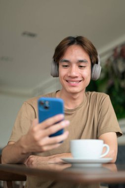 Kahverengi tişörtlü genç Taylandlı öğrenci sosyal medya, uzaktan iletişim ve dijital yaşam tarzı gibi modern bir kafede boynuna kulaklık takıyor..