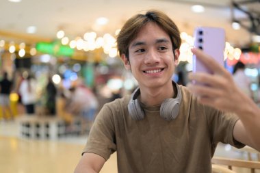 Kahverengi tişörtlü genç Taylandlı öğrenci sosyal medya, uzaktan iletişim ve dijital yaşam tarzı gibi modern bir kafede boynuna kulaklık takıyor..