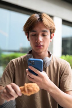 Fast food, aperatif ve yemek konsepti için açık bir masada kızarmış tavuk budu yemeye hazır kulaklıklı, kahverengi tişörtlü genç Asyalı öğrenci.