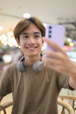 Kahverengi tişörtlü genç Taylandlı öğrenci sosyal medya, uzaktan iletişim ve dijital yaşam tarzı gibi modern bir kafede boynuna kulaklık takıyor..