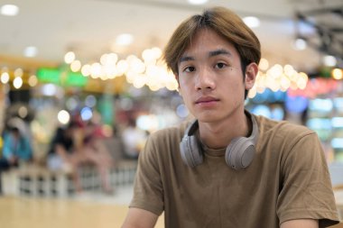 Kahverengi tişörtlü genç Taylandlı öğrenci sosyal medya, uzaktan iletişim ve dijital yaşam tarzı gibi modern bir kafede boynuna kulaklık takıyor..