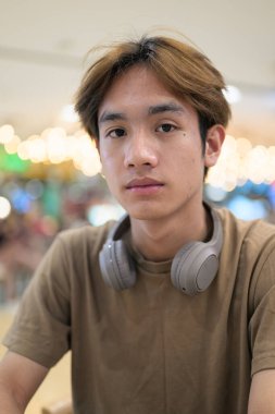 Kahverengi tişörtlü genç Taylandlı öğrenci sosyal medya, uzaktan iletişim ve dijital yaşam tarzı gibi modern bir kafede boynuna kulaklık takıyor..