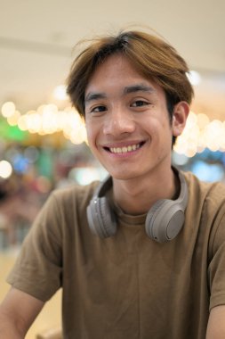 Kahverengi tişörtlü genç Taylandlı öğrenci sosyal medya, uzaktan iletişim ve dijital yaşam tarzı gibi modern bir kafede boynuna kulaklık takıyor..