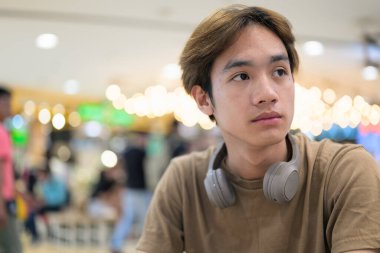 Kahverengi tişörtlü genç Taylandlı öğrenci sosyal medya, uzaktan iletişim ve dijital yaşam tarzı gibi modern bir kafede boynuna kulaklık takıyor..