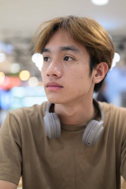 Kahverengi tişörtlü genç Taylandlı öğrenci sosyal medya, uzaktan iletişim ve dijital yaşam tarzı gibi modern bir kafede boynuna kulaklık takıyor..