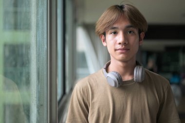 Genç yakışıklı ve zayıf Asyalı Taylandlı adam spor salonunda pencerenin yanında kulaklık takıyor.