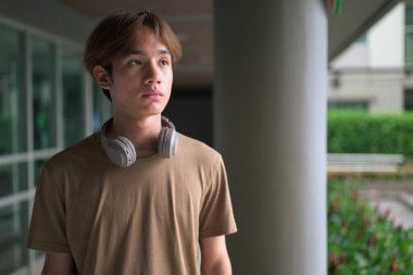 Kulaklıklı yakışıklı Asyalı genç Taylandlı. Düşünüp başka tarafa bakarken modern yaşam tarzının tadını çıkarıyor.