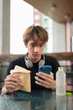 Mutlu Asyalı genç Taylandlı öğrenci dışarıda öğle yemeği için sandviçini yerken oturup telefonla konuşuyor.