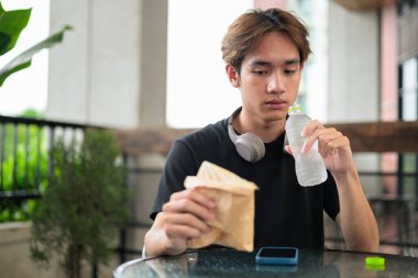Mutlu Asyalı genç Taylandlı öğrenci sandviç yiyor ve öğle yemeği için açık havada su içiyor.