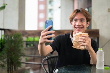 Mutlu Asyalı genç Taylandlı öğrenci dışarıda öğle yemeği için sandviçini yerken oturup telefonla konuşuyor.