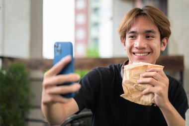 Mutlu Asyalı genç Taylandlı öğrenci dışarıda öğle yemeği için sandviçini yerken oturup telefonla konuşuyor.