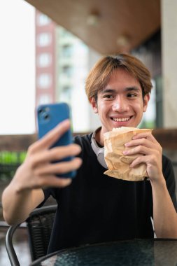 Mutlu Asyalı genç Taylandlı öğrenci dışarıda öğle yemeği için sandviçini yerken oturup telefonla konuşuyor.