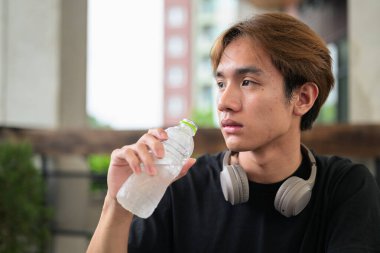 Genç adam dışarıda otururken şişeden su içiyor ve kulaklıkları boynunda.