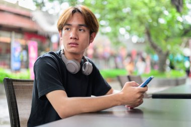 Taylandlı genç bir adamın portresi. Sıradan tarzda oturuyor ve yazın açık havada telefon kullanıyor. Çeşitli kavramlar ve kullanımlar için kendine güvenen, rahat ve otantik görünüm