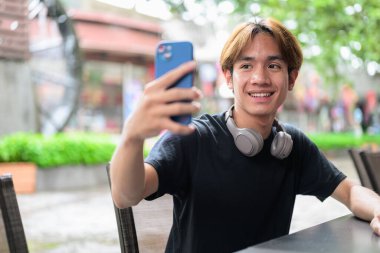 Taylandlı genç bir adamın portresi. Sıradan tarzda oturuyor ve yazın açık havada telefon kullanıyor. Çeşitli kavramlar ve kullanımlar için kendine güvenen, rahat ve otantik görünüm