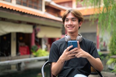 Genç Taylandlı bir adam geleneksel Çin bahçe pazarında akıllı telefon kullanarak dışarıda oturuyor. Çeşitli kavramlar ve kullanımlar için kendine güvenen, rahat ve otantik görünüm