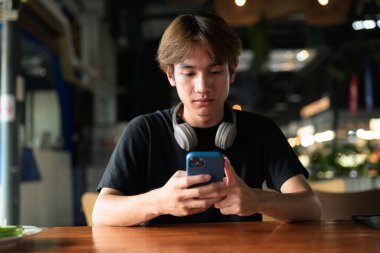 Gündüz otururken cep telefonu kullanan Taylandlı genç bir adamın portresi. Çeşitli kavramlar ve kullanımlar için kendine güvenen, rahat ve otantik görünüm