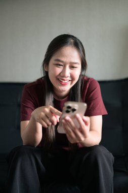Genç Taylandlı kadın akıllı telefon kullanarak evdeki kanepede oturuyor. Teknolojiyi, rahatlamayı ve günlük iletişimi gösteren modern yaşam tarzı portresi.