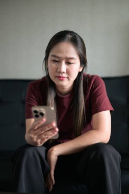 Genç Taylandlı kadın akıllı telefon kullanarak evdeki kanepede oturuyor. Teknolojiyi, rahatlamayı ve günlük iletişimi gösteren modern yaşam tarzı portresi.