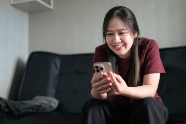 Genç Taylandlı kadın akıllı telefon kullanarak evdeki kanepede oturuyor. Teknolojiyi, rahatlamayı ve günlük iletişimi gösteren modern yaşam tarzı portresi.