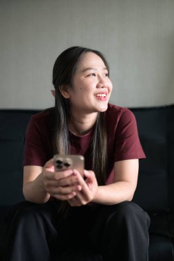 Genç Taylandlı kadın akıllı telefon kullanarak evdeki kanepede oturuyor. Teknolojiyi, rahatlamayı ve günlük iletişimi gösteren modern yaşam tarzı portresi.