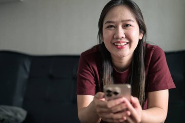 Genç Taylandlı kadın akıllı telefon kullanarak evdeki kanepede oturuyor. Teknolojiyi, rahatlamayı ve günlük iletişimi gösteren modern yaşam tarzı portresi.