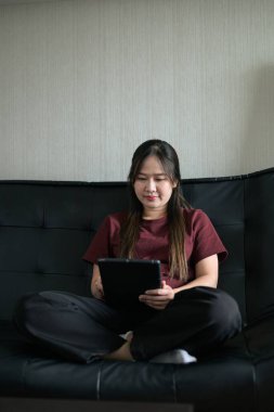 Genç Taylandlı kadın evdeki dijital tablet bilgisayarını kullanarak kanepede oturuyor. Gelişigüzel teknoloji kullanımını, çevrimiçi taramayı ve rahatlamayı gösteren modern yaşam tarzı portresi.