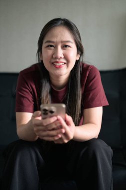 Genç Taylandlı kadın akıllı telefon kullanarak evdeki kanepede oturuyor. Teknolojiyi, rahatlamayı ve günlük iletişimi gösteren modern yaşam tarzı portresi.