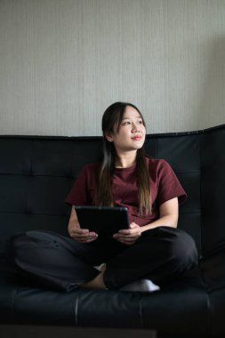 Genç Taylandlı kadın evdeki dijital tablet bilgisayarını kullanarak kanepede oturuyor. Gelişigüzel teknoloji kullanımını, çevrimiçi taramayı ve rahatlamayı gösteren modern yaşam tarzı portresi.