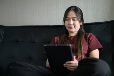 Genç Taylandlı kadın evdeki dijital tablet bilgisayarını kullanarak kanepede oturuyor. Gelişigüzel teknoloji kullanımını, çevrimiçi taramayı ve rahatlamayı gösteren modern yaşam tarzı portresi.