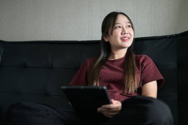 Genç Taylandlı kadın evdeki dijital tablet bilgisayarını kullanarak kanepede oturuyor. Gelişigüzel teknoloji kullanımını, çevrimiçi taramayı ve rahatlamayı gösteren modern yaşam tarzı portresi.