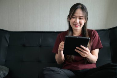 Genç Taylandlı kadın evdeki dijital tablet bilgisayarını kullanarak kanepede oturuyor. Gelişigüzel teknoloji kullanımını, çevrimiçi taramayı ve rahatlamayı gösteren modern yaşam tarzı portresi.