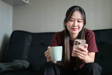 Genç Taylandlı kadın evinde kanepede oturuyor. Akıllı telefon kullanıyor ve elinde kahve fincanı tutuyor. Teknolojiyi, rahatlamayı ve günlük iletişimi gösteren modern yaşam tarzı portresi.