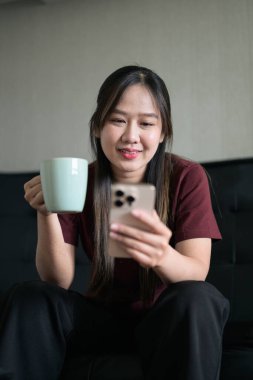 Genç Taylandlı kadın evinde kanepede oturuyor. Akıllı telefon kullanıyor ve elinde kahve fincanı tutuyor. Teknolojiyi, rahatlamayı ve günlük iletişimi gösteren modern yaşam tarzı portresi.