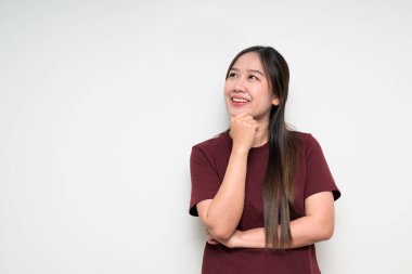 Asyalı genç bir kadının düşünme ve gülümseme portresi. Beyaz arka planda el ele tutuşup günlük tişört giyerken.