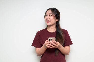 Beyaz arka plan üzerinde düşünürken ve gülümserken cep telefonu kullanan Asyalı genç bir kadının stüdyo portresi.