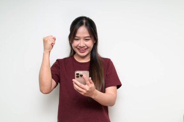 Beyaz arka planda cep telefonu kullanan, kolları havada, rahat bir tişört giyen Asyalı bir kadının stüdyo portresi.