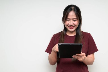 Beyaz arka planda gülümseyen Asyalı genç bir kadının günlük tişört giyip dijital tablet bilgisayar kullanan bir stüdyo portresi.