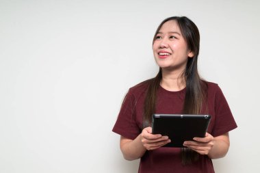 Beyaz arka planda, sıradan tişörtler giyen ve düşünürken ve gülümserken dijital tablet bilgisayar kullanan Asyalı genç bir kadının stüdyo portresi.