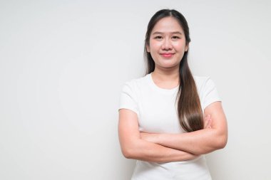 Kolları düz arkaplanda çaprazlanmış beyaz tişört giyen Asyalı genç bir kadının stüdyo portresi.