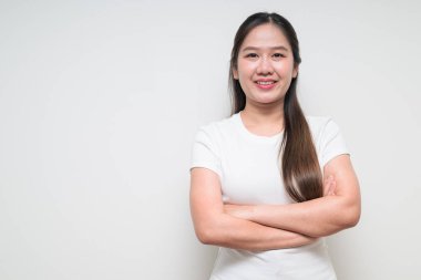 Kolları düz arkaplanda çaprazlanmış beyaz tişört giyen Asyalı genç bir kadının stüdyo portresi.