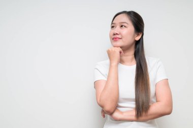 Sıradan beyaz tişört giyerek arka planda düşünen ve planlayan Asyalı genç bir kadının stüdyo portresi.