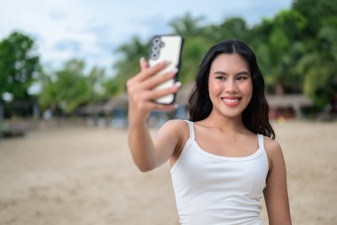 Genç ve güzel Asyalı bir kadın yaz tatilinde sahilde telefonuyla selfie çekiyor.