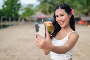 Genç ve güzel Asyalı bir kadın yaz tatilinde elinde hindistan cevizi tutarken sahilde telefonuyla selfie çekiyor.