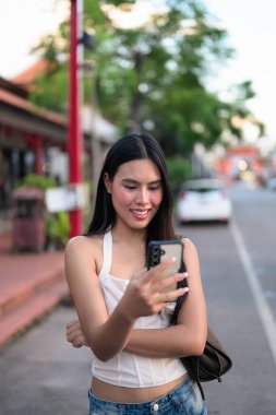Genç ve güzel Asyalı bir kadın yazın gülümseyerek ve telefonla caddede taksi bekliyor.