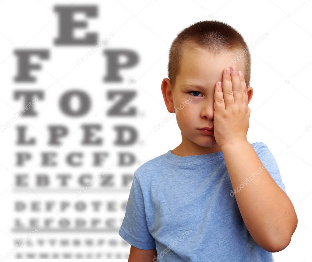 Little boy covering one eye with palm for eyesight exam with blurry eye chart behind