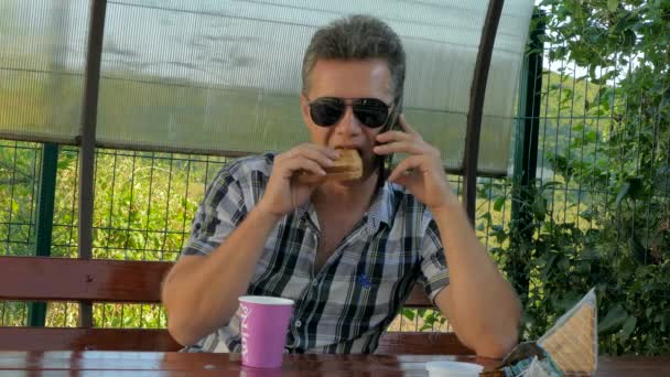 Egy ember szemüveg a pavilonban eszik egy kávét szendvicset — Stock videók