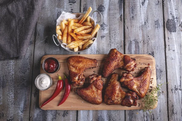 Grilled chicken wings, french fries, white and red sauce nuts on a wooden surface. Bottle and glass of beer