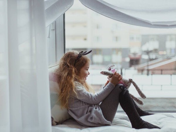 Pretty girl sitting near window and playing with soft toy, intdoor