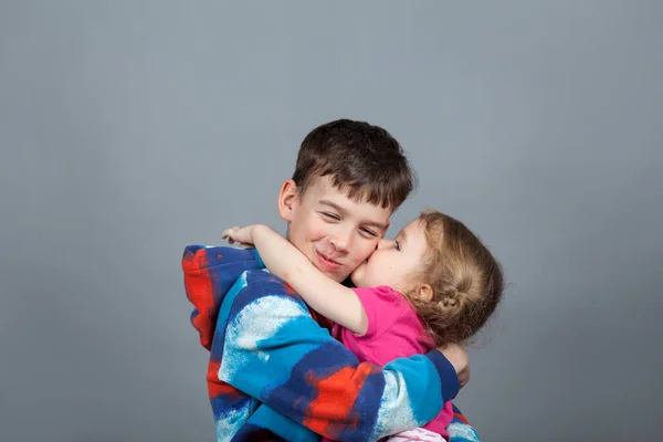 Sister hugs older brother and kisses him on the cheek. Studio gray background